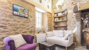 Ein Zimmer mit Steinwänden, einem weißen Sofa, einem violetten Sessel, einem Holztisch und einem Bücherregal.