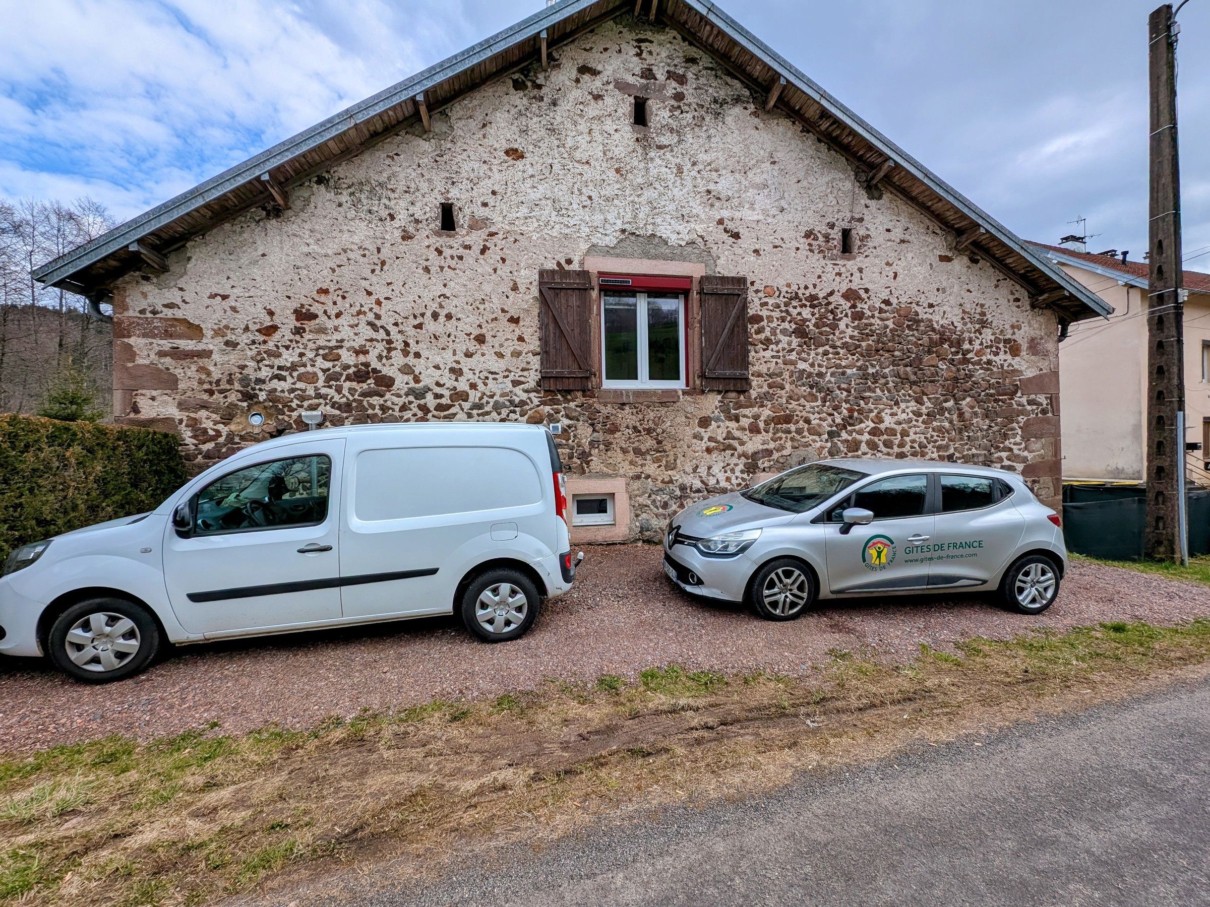 Deux voitures sont garées devant un bâtiment rustique en pierre, doté d’un toit en pente et d’une petite fenêtre avec des volets.