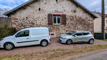 Deux voitures sont garées devant un bâtiment rustique en pierre, doté d’un toit en pente et d’une petite fenêtre avec des volets.