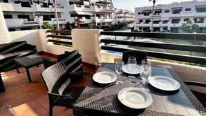 Et udendørs spiseområde med et bord dækket til to, omgivet af fletstole, på en terrasse med udsigt til boliger.