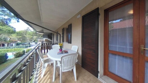 A balcony with a table and chairs, a view of greenery, and a large wooden door. - Spacious Apartment With two Balconies (Lignano Sabbiadoro)