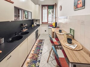 A narrow kitchen with a black countertop, a window with curtains, a tiled backsplash, and a dining area with a table and chairs.