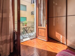 Una stanza con pavimento in legno, una porta scorrevole che dà su un balcone con tavolo e sedie, e un frigorifero.