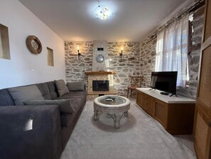 A living room with a stone wall, a fireplace, a sofa, a coffee table, a television, and a cabinet. - White Stone Apartments - Ground Floor (Sithonia)