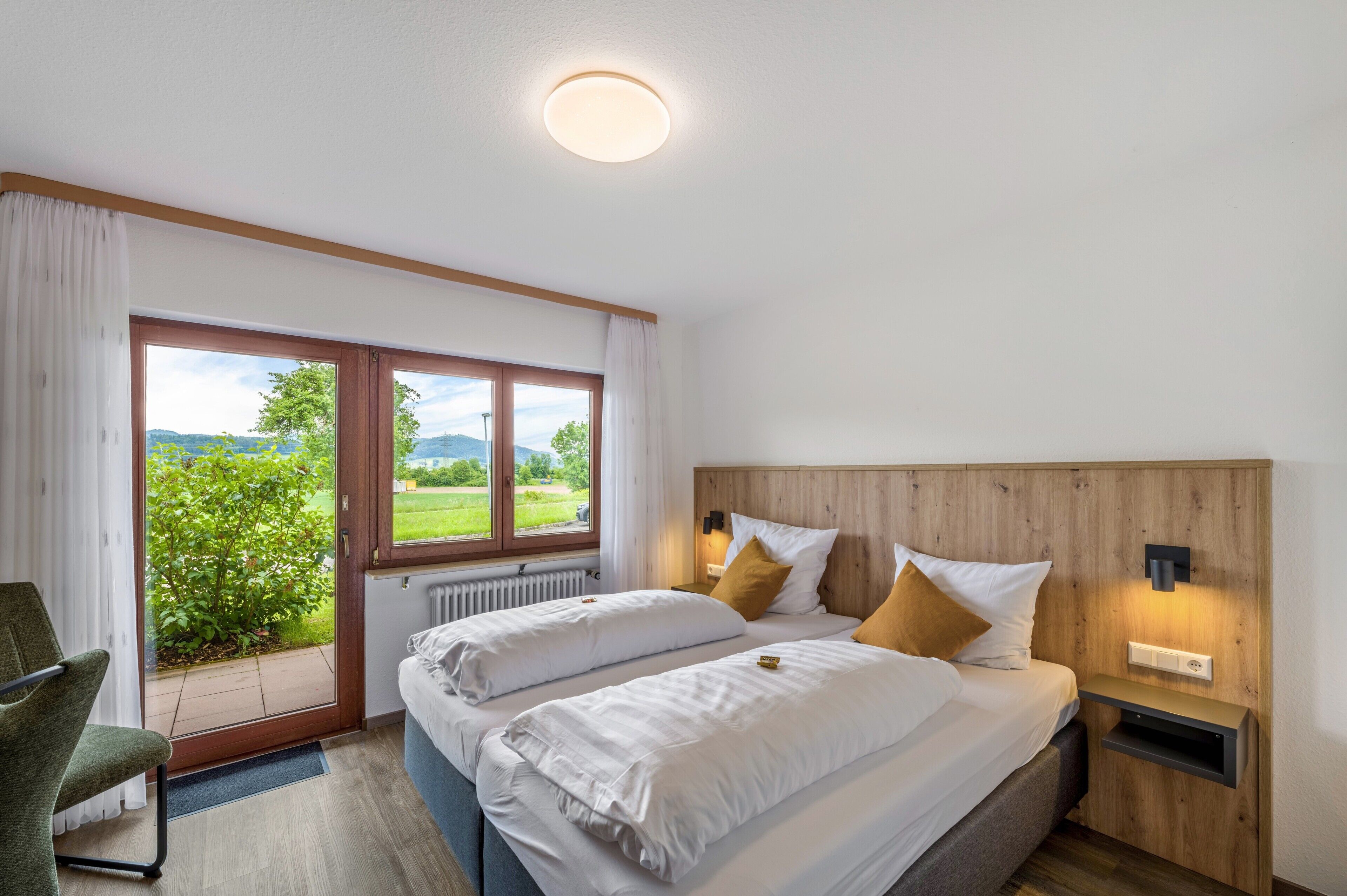 A hotel room with two beds, a view of greenery, a wooden headboard, and a small bedside table with a lamp.