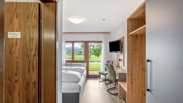 Quarto de hotel com cama, escrivaninha, cadeira, televisão e janela com vista para área verde.