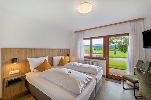 Ein Hotelzimmer mit einem Holzkopfteil, zwei Betten mit weißer Bettwäsche, einem grünen Sessel, einem Fernseher und einem großen Fenster mit Blick auf Grünflächen und eine Straße.