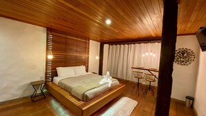 Un dormitorio con techo de madera, una cama, una mesita de noche, un escritorio con dos sillas, una ventana con cortinas y un aplique de pared.