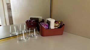 Habitación de hotel con una mesa, tres vasos vacíos, una caja de pañuelos y una cesta con aperitivos y una botella.