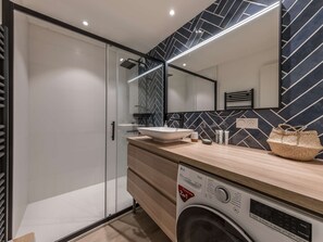 Ein modernes Badezimmer mit Duschkabine aus Glas, einem Waschbecken mit Vessel-Becken, einer Waschmaschine und einem Spiegel mit geometrischer Rückwand.