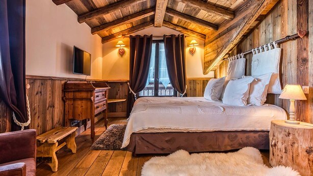A rustic bedroom with a wooden ceiling, a bed with white linens, a wooden dresser, a bench, and a fur rug. - Cozy 2BR Apartment in Courchevel 1850 – Center, Close to Slopes, WiFi, Hammam, Ski Locker (Courchevel)