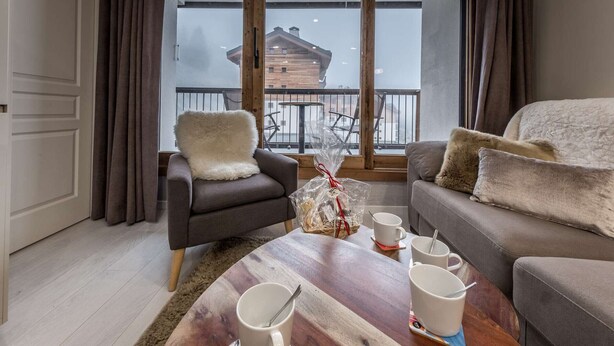 Un salón acogedor con sofá, sillón y mesa de centro, con vistas a un edificio de madera a través de grandes ventanales.