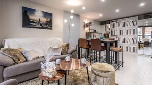 Una sala de estar moderna con sofá, mesa de centro y reposapiés, una zona de comedor con sillas y mesa, una estantería y un cuadro en la pared.