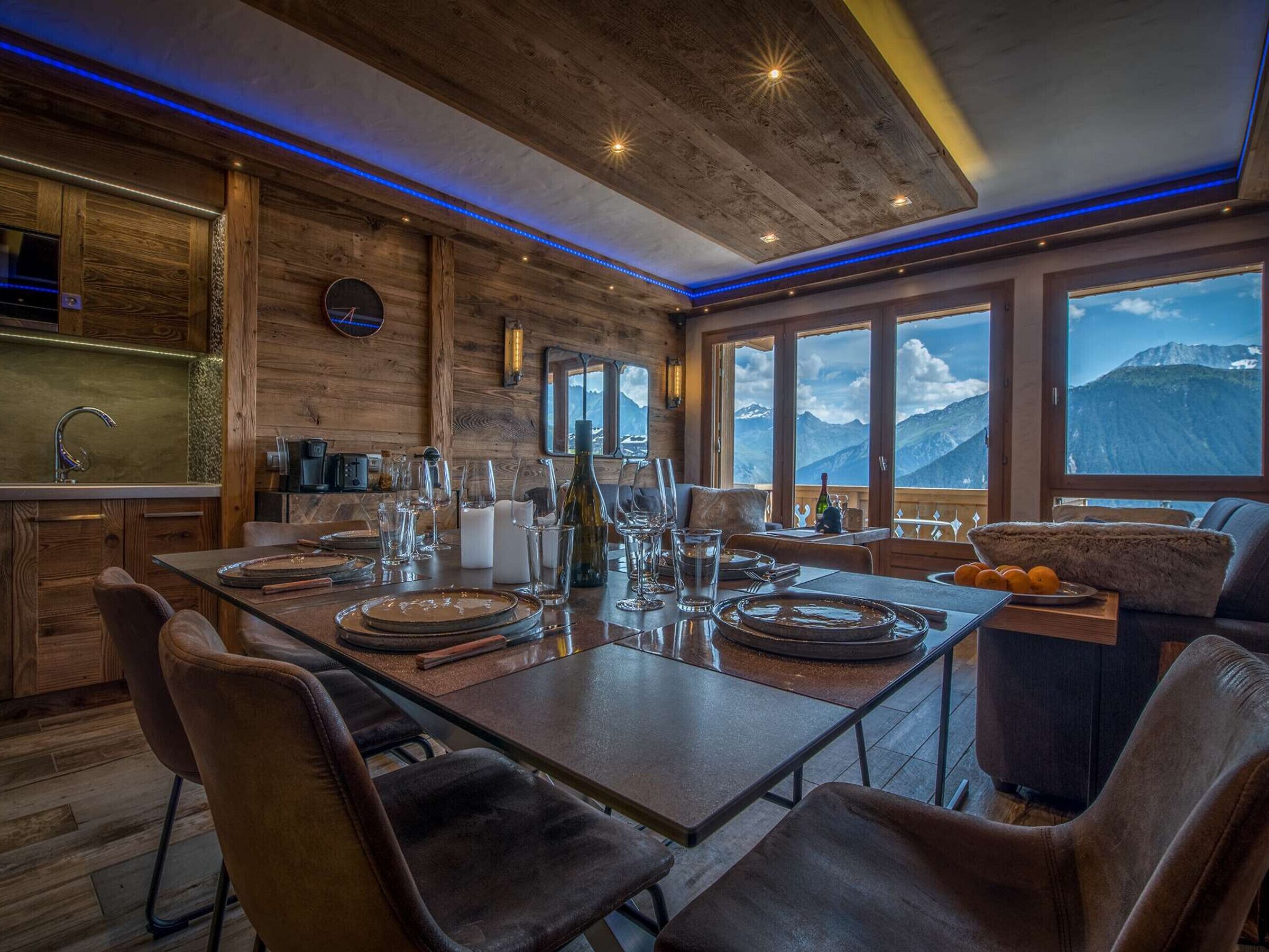A dining area with a wooden table set for four, a view of mountains through large windows, and a modern kitchen with wooden accents.