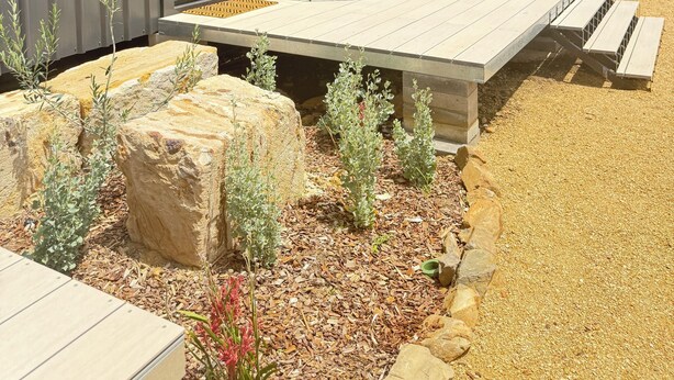 A modern outdoor deck with a sofa and grill, surrounded by rocks and plants.