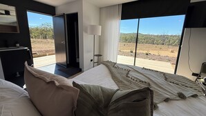 A modern bedroom with a large bed, a bedside table, a lamp, and a view of a dry landscape.