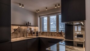 Une cuisine moderne avec des meubles de couleur sombre, un plan de travail en marbre et une grande fenêtre donnant sur des bâtiments enneigés.