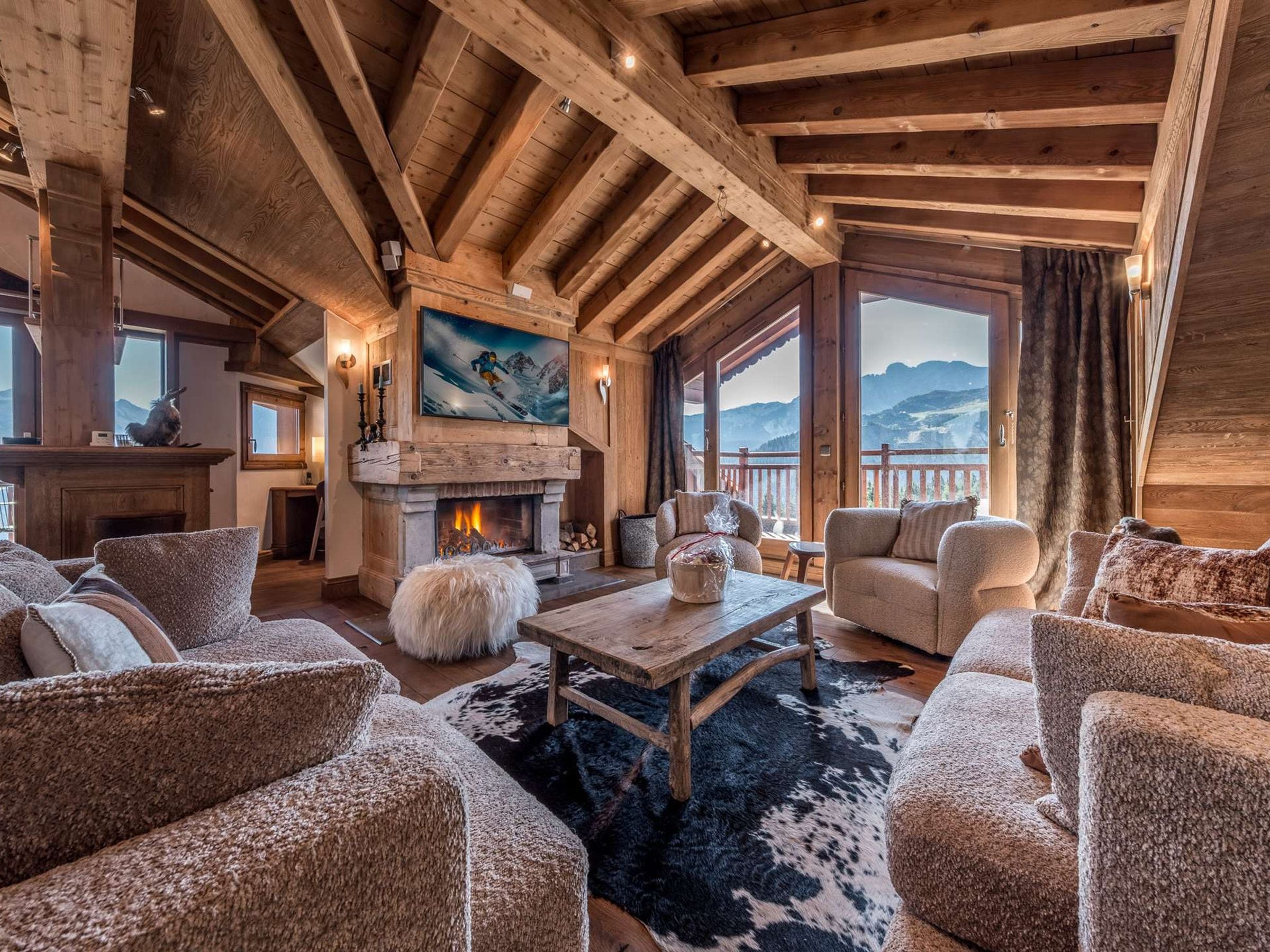 A cozy lodge interior with a fireplace, wooden beams, plush seating, and a scenic mountain view.