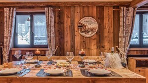 Mesa de jantar de madeira posta com pratos, copos e velas, com parede e janelas de madeira ao fundo.
