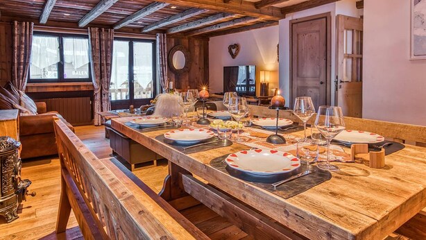 Uma mesa de jantar de madeira posta com pratos, copos e talheres, um sofá e uma lareira.