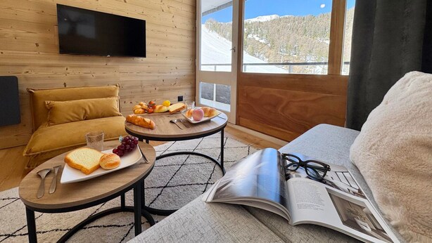 Una camera accogliente con parete in legno, TV a schermo piatto, tavolo da pranzo con colazione, divano e finestra con vista innevata.