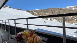 Un balcone con un tavolo su cui ci sono pane, uva e un bicchiere d'acqua, con vista su una pista da sci innevata.