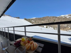 Ein Balkon mit Tisch, auf dem Brot, Trauben und ein Glas Wasser stehen, mit Blick auf eine verschneite Skipiste.