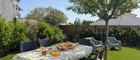 An outdoor dining area with a table set for breakfast, surrounded by greenery and a tree.