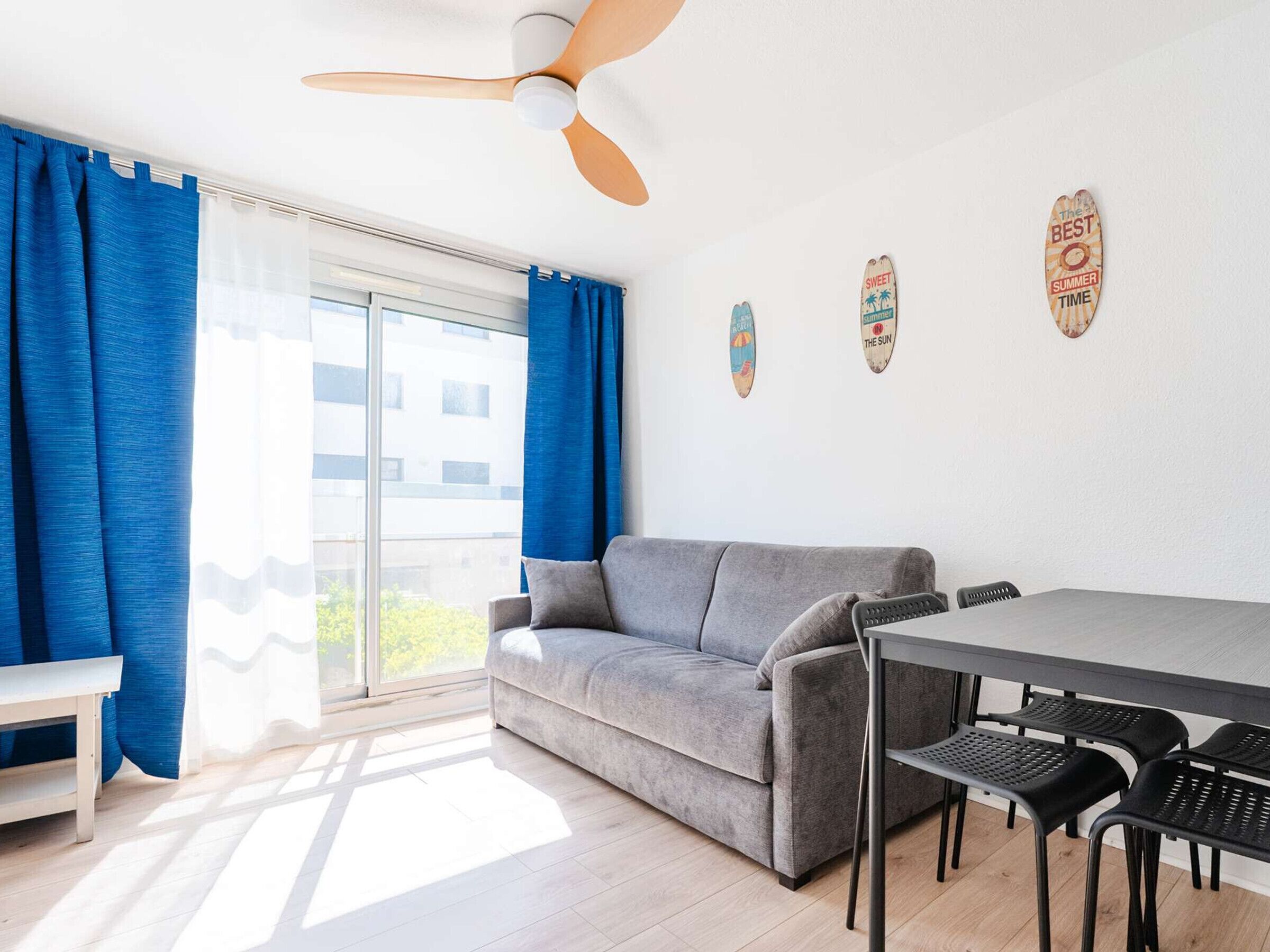 Uma sala de estar moderna com um sofá cinza, uma mesa de jantar com quatro cadeiras, um ventilador de teto e uma janela grande com cortinas azuis.