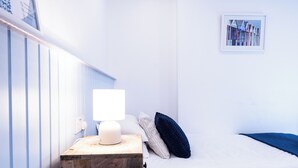 Un dormitorio con una mesita de noche de madera, una lámpara blanca, una cama con ropa de cama blanca, un almohacer azul y un cuadro en la pared.