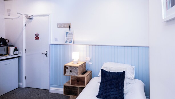 Una habitación de hotel con cama, mesita de noche con lámpara, un pequeño armario de madera, escritorio con tetera y un cartel de 'No Smoking'.