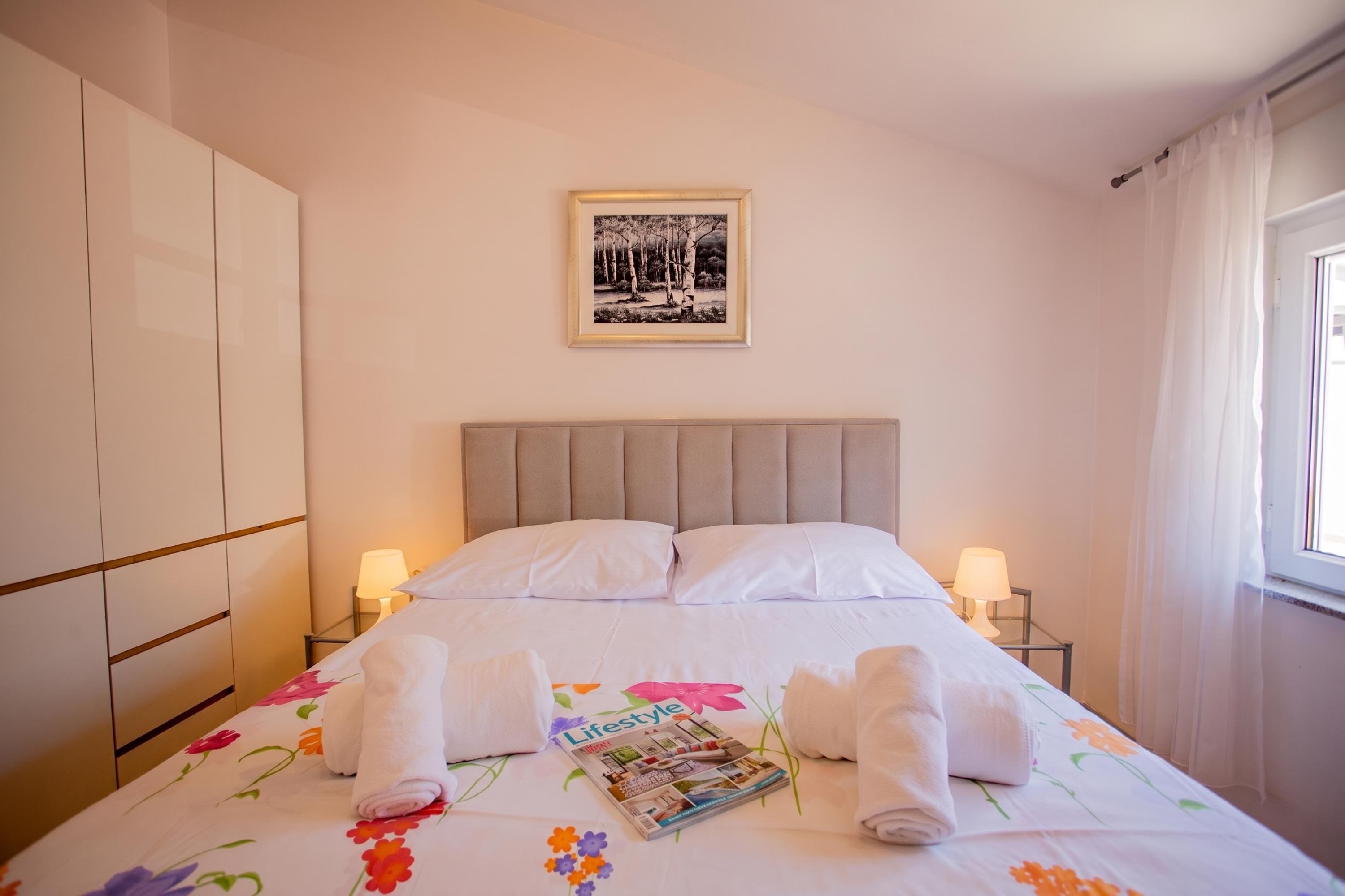 Un lit bien fait, recouvert d’une couvre-lit à motifs floraux, deux lampes de chevet, un tableau encadré au mur et un magazine posé sur le lit.