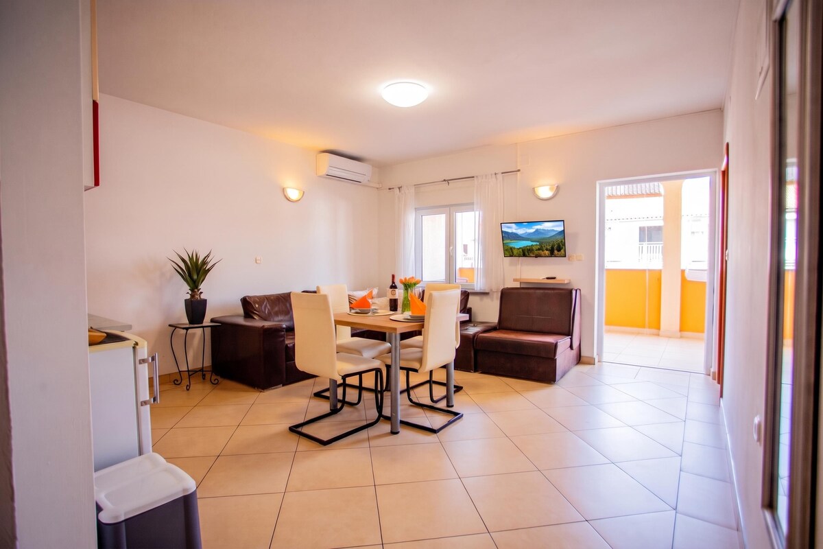 A modern living room with a dining area, a flat-screen TV mounted on the wall, and a sliding glass door leading to an outdoor balcony.