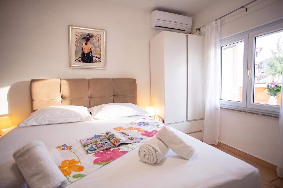 A hotel room with a bed, a framed picture on the wall, an air conditioner, and a window with curtains.