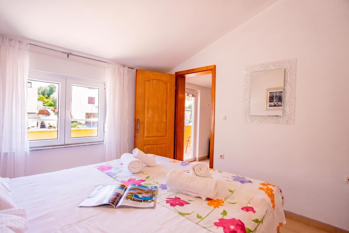 A hotel room with a bed, a window with curtains, a door, and a wall-mounted picture.