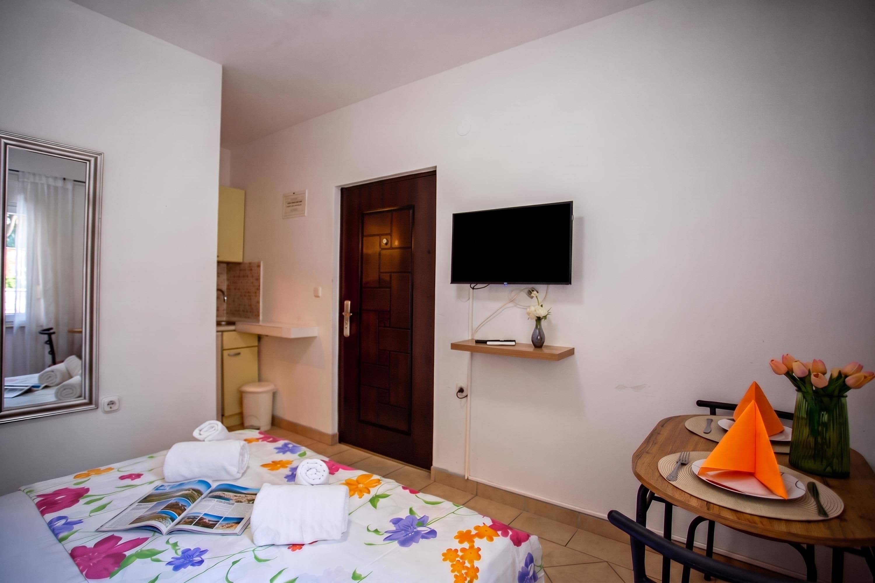 Ein Hotelzimmer mit einem Bett, einem an der Wand befestigten Fernseher, einem kleinen Tisch mit Blumenvase und einer Tür zu einem weiteren Raum.