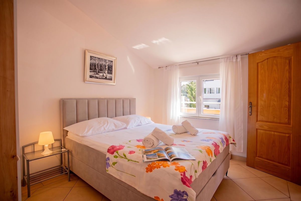 A bedroom with a bed, a bedside table with a lamp, a framed picture on the wall, and a window with curtains.