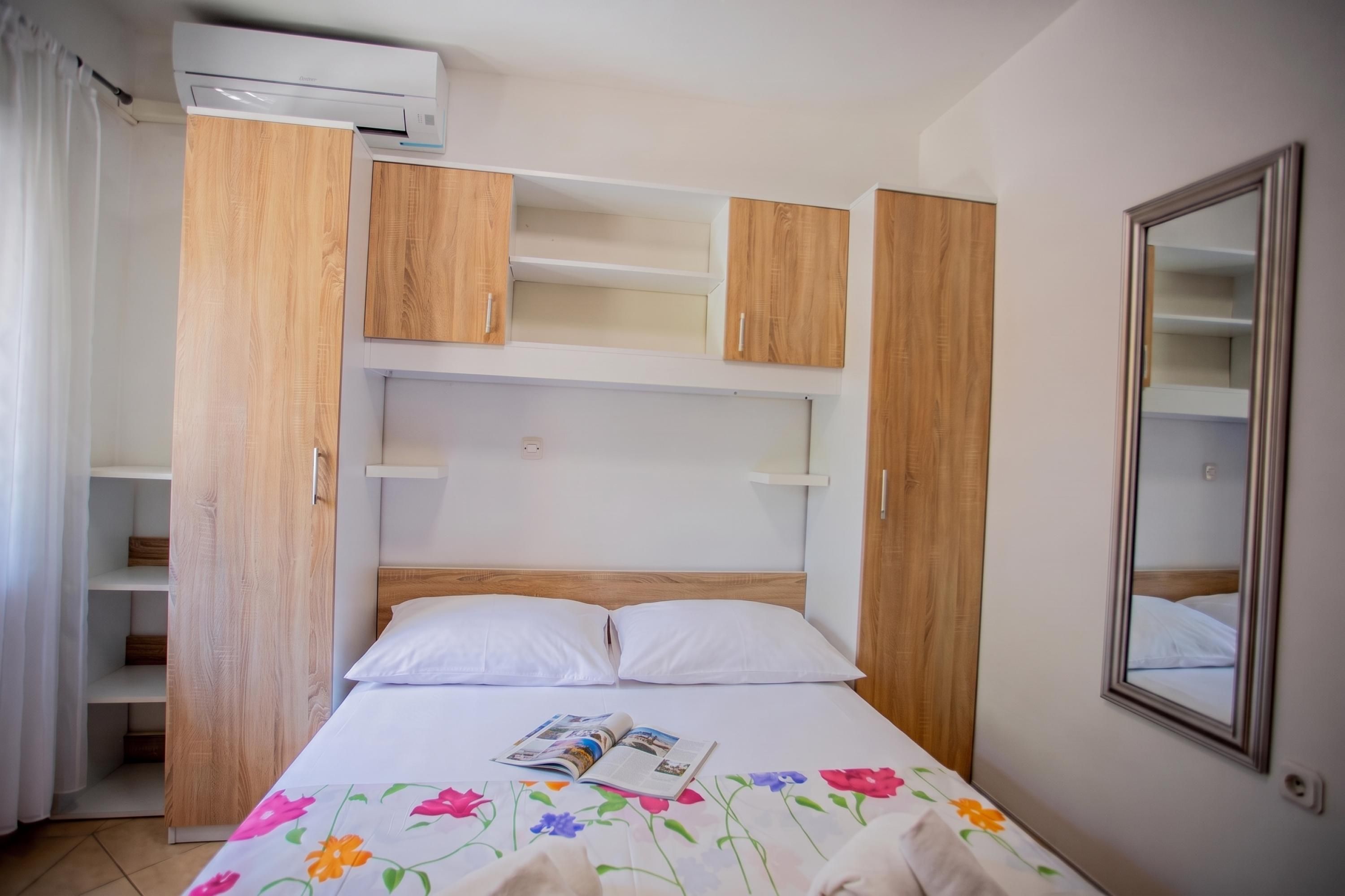 A bedroom with a bed, a wooden wardrobe, a mirror, and a built-in air conditioning unit.