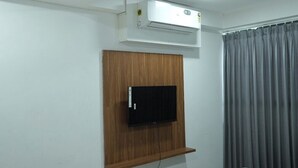A room with a mounted air conditioner, a flat-screen TV on a wooden panel, and a grey curtain. - HOTEL KRISHNA INN (Ahmedabad)