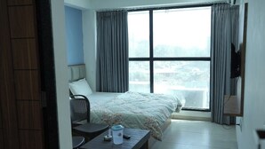 A hotel room with a bed, a chair, a small table, a TV, and a window with curtains. - HOTEL KRISHNA INN (Ahmedabad)