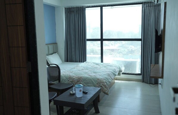 A hotel room with a bed, a chair, a small table, a TV, and a window with curtains. - HOTEL KRISHNA INN (Ahmedabad)