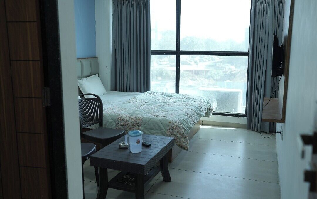 A bedroom with a bed, a small table with a cup and remote, a chair, and a window with curtains.