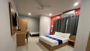 A hotel room with a bed, a desk, a chair, a television, an air conditioner, and a ceiling fan. - Surbhi Grant (Khatoo)