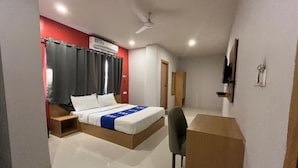 A hotel room with a bed, a desk, a chair, a TV, and a ceiling fan. - Surbhi Grant (Khatoo)