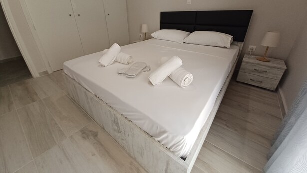A neatly made bed with white linens, a pair of slippers, and rolled towels. There is a bedside table with a lamp and a small drawer. The room has a tiled floor and a wardrobe with a dark headboard. - Marina Zeas Hidden Gem (Pireas)