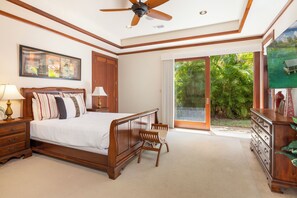Ein Schlafzimmer mit einem Bett, einem Nachttisch, einer Kommode, einem Sessel, einem Deckenventilator und einem großen Fenster mit Blick auf Grünflächen.