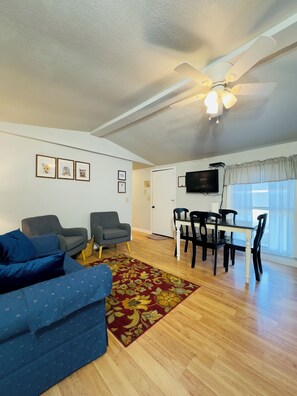 A living room with a ceiling fan, a TV, a dining table with chairs, and a sofa with cushions. - Cabin 7 2-bedroom house along the Gasconade River on a lovely Campground Resort (Richland)