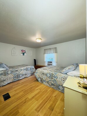 Ein Zimmer mit zwei Betten, Holzboden, einem Fenster mit Vorhängen, einem Heizkörper und einer Deckenleuchte.