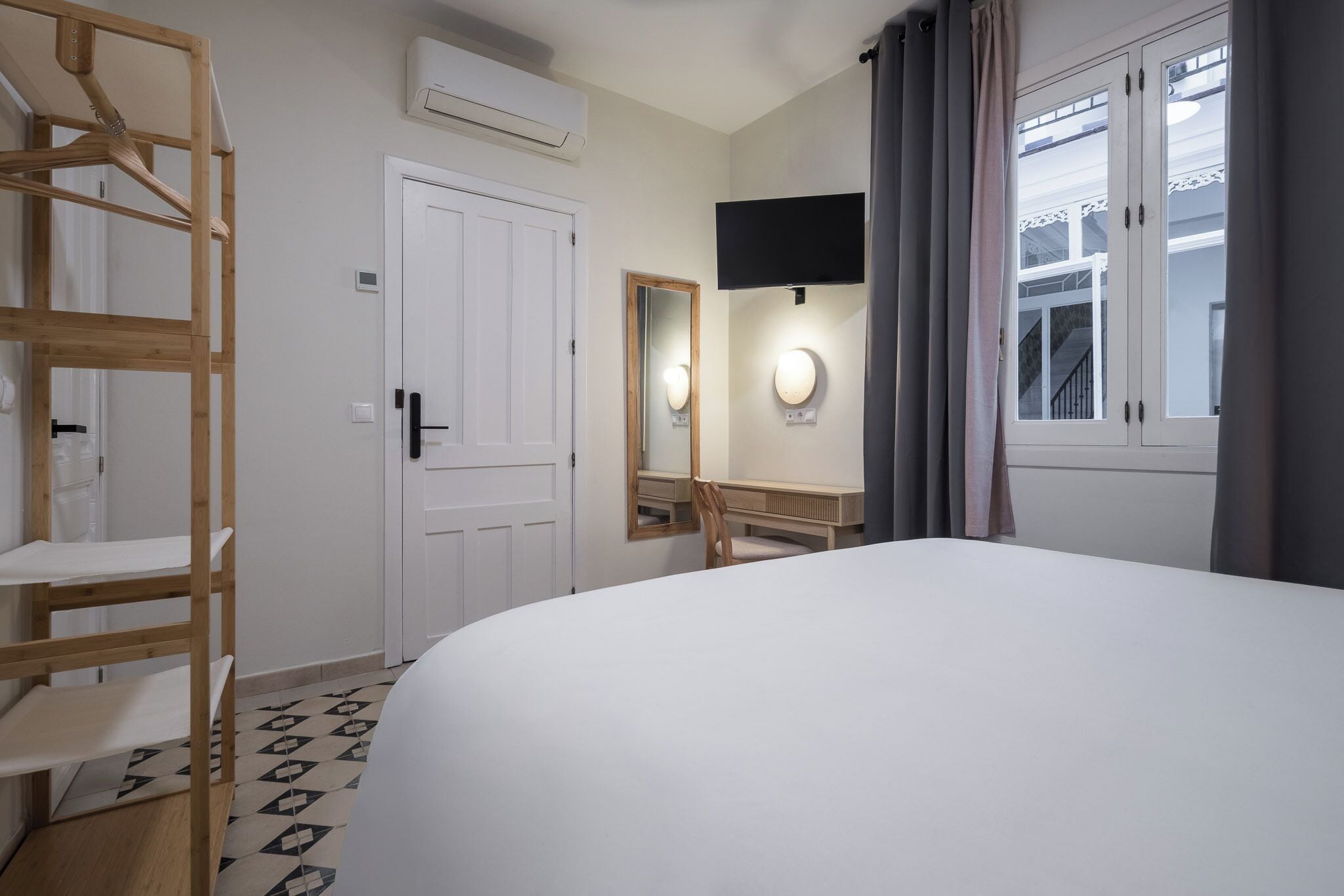 Ein Hotelzimmer mit einem Bett, einer Holzhockerreihe, einem Spiegel, einem Fernseher und einem Fenster mit Vorhängen.