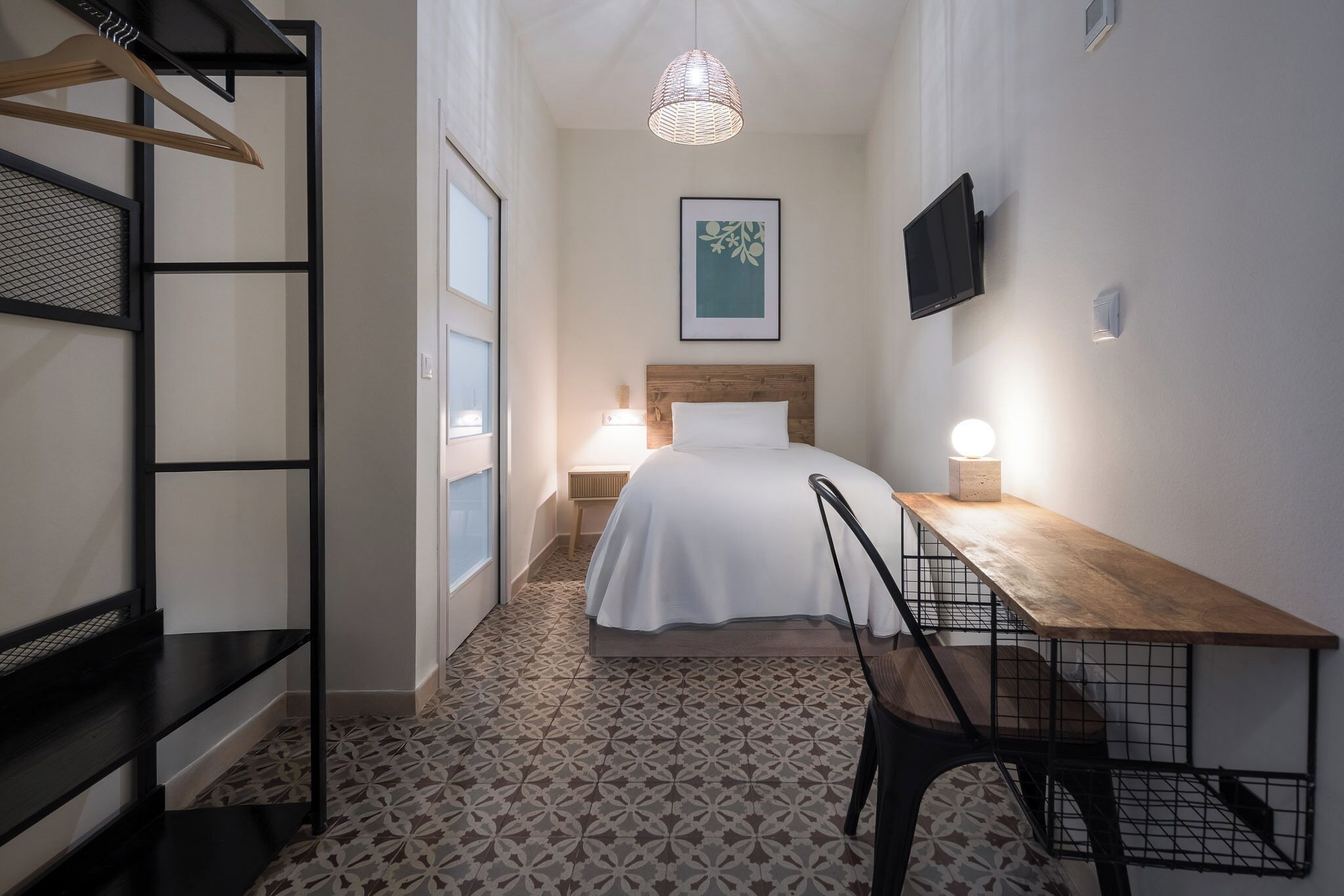 Habitación de hotel estrecha con cama, mesita de noche, silla y lámpara colgante.